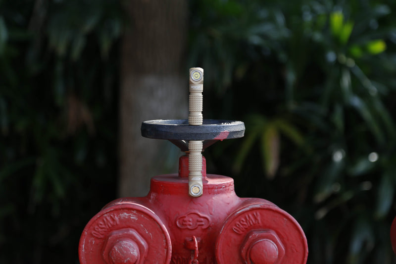 A close-up of a red outdoor water valve with two connections, surrounded by blurred green foliage in the background, illuminated by the Manker E03H III flashlight with 1200 lumens that highlights every detail.