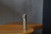 A small, brown Manker E03H III right-angle flashlight is positioned upright on a wooden table with a softly blurred background.