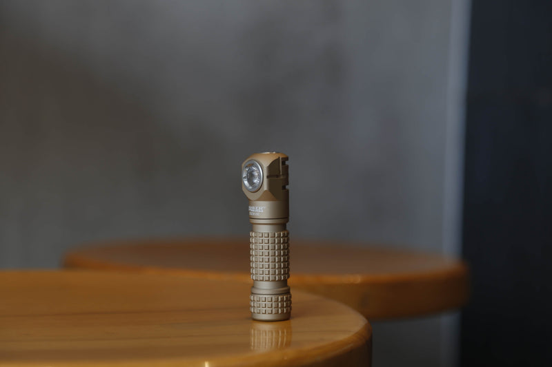 A small, brown Manker E03H III right-angle flashlight is positioned upright on a wooden table with a softly blurred background.