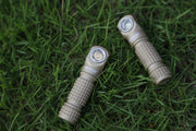 Two tan-colored flashlights, including the Manker E03H III headlamp, lie on green grass, positioned parallel to each other with their lenses facing upward.