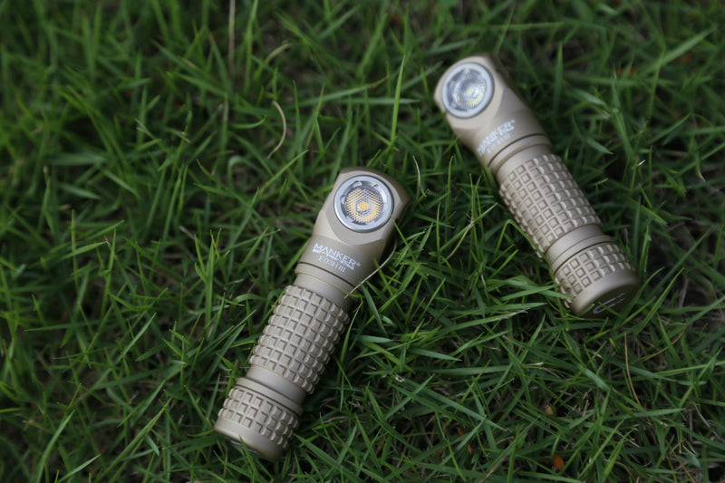 Two tan-colored flashlights, including the Manker E03H III headlamp, lie on green grass, positioned parallel to each other with their lenses facing upward.