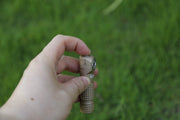 A hand holds a tan Manker E03H III flashlight with a side button, pictured against a blurred green grass background.