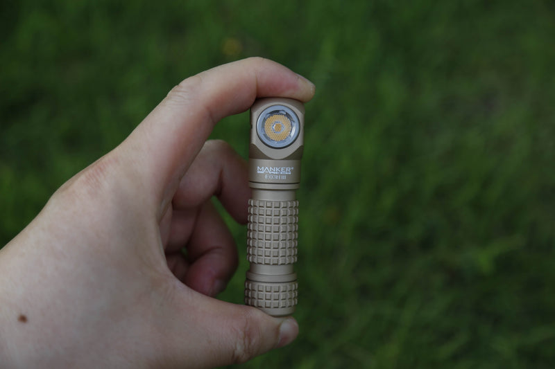A hand holds a compact, tan-colored Manker E05 II flashlight with a textured grip against a blurred green background, highlighting its design alongside the Manker E03H III, renowned for its outstanding brightness of up to 1200 lumens.