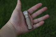 A person holds a compact, tan Manker E03H III flashlight with a textured grip in their open palm against a green grass background, highlighting its impressive 1200-lumen output.