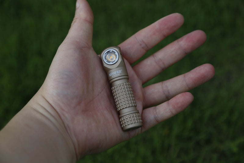 A person holds a compact, tan Manker E03H III flashlight with a textured grip in their open palm against a green grass background, highlighting its impressive 1200-lumen output.