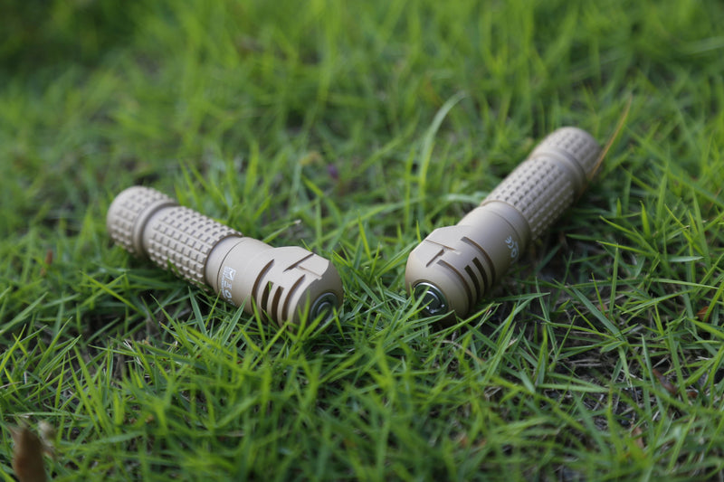 Two tan Manker E03H III flashlights with textured grips, each known for its 1200 lumens output and reputation as the brightest headlamp, are lying on green grass, positioned parallel to each other with the lenses facing outward.