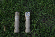 Two Manker E03H III flashlights with tan-colored, textured grips are lying side by side on green grass—one displaying the LED front and the other showing its marked side. The Manker E03H III stands out for its remarkable brightness, reaching up to 1200 lumens, making it a top contender among the brightest headlamps in its class.