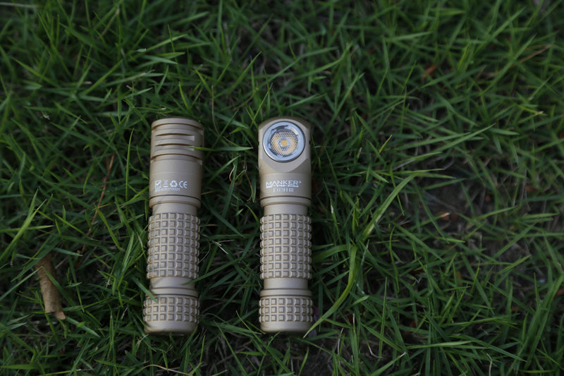 Two Manker E03H III flashlights with tan-colored, textured grips are lying side by side on green grass—one displaying the LED front and the other showing its marked side. The Manker E03H III stands out for its remarkable brightness, reaching up to 1200 lumens, making it a top contender among the brightest headlamps in its class.