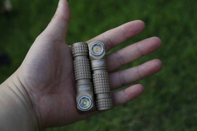 A hand holds two small, brown, textured flashlights—each bearing the Manker branding—with one model identifiable as the Manker E03H III, which boasts a powerful 1200-lumen output. Lush green grass forms the background.