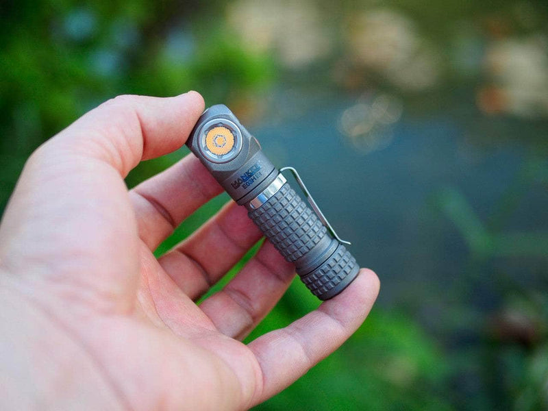 A hand holds the Manker E03H III - Titanium, a compact gray flashlight with a side button and textured grip, ideal for everyday carry and outdoor adventures near greenery and water.