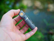 A hand holds the Manker E03H III - Titanium, a compact gray right-angle titanium flashlight with a textured grip and pocket clip, perfect for everyday carry, outdoors amid greenery.