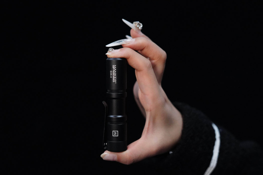 A person's hand holding a Manker E05 II flashlight on a black background.