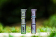 Two Manker E05 II Ti - Colorful MAO Limited Edition flashlights stand upright outdoors side by side, ideal for everyday carry, with a blurred green background.