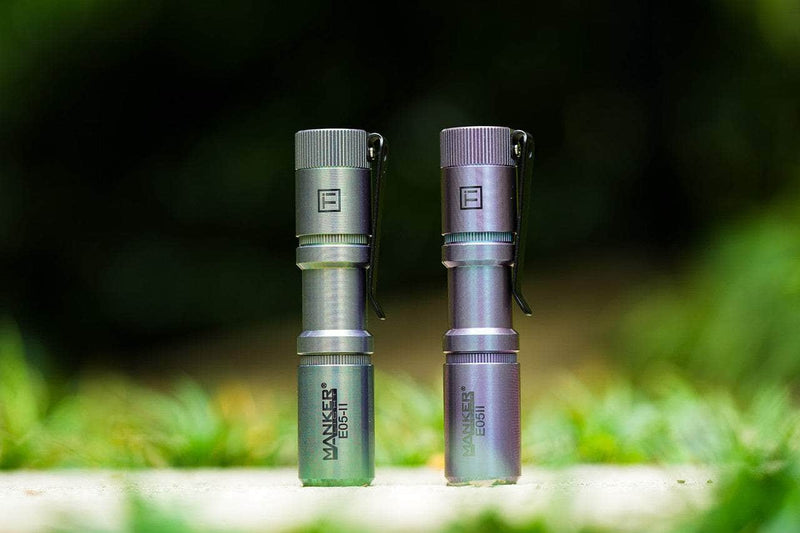 Two Manker E05 II Ti - Colorful MAO Limited Edition flashlights stand upright outdoors side by side, ideal for everyday carry, with a blurred green background.