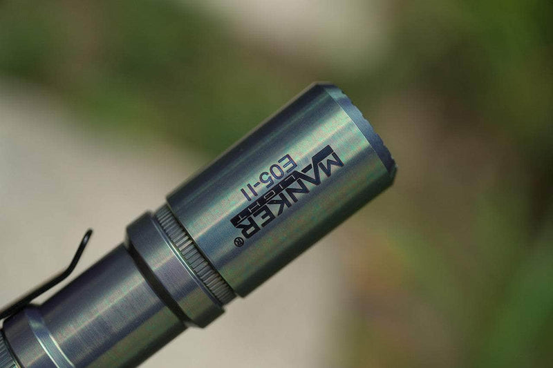 Close-up of the limited edition Manker E05 II Ti - Colorful MAO flashlight, delivering 1300 lumens, set against a blurred green and white background—ideal for everyday carry.