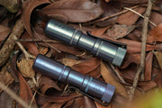 Two Manker E05 II Ti - Colorful MAO Limited Edition titanium flashlights, ideal for everyday carry, lie on dry brown leaves and twigs outdoors.