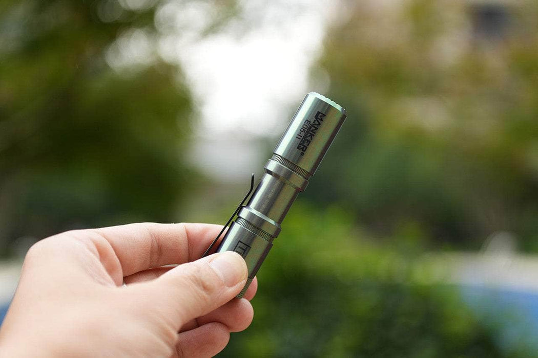 A hand holds the Manker E05 II Ti - Colorful MAO Limited Edition titanium flashlight outdoors, with blurred greenery in the background—an ideal EDC tool delivering reliable illumination up to 1300 lumens.
