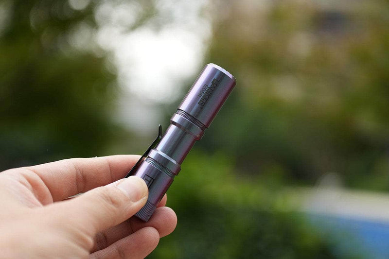 A hand holds the Manker E05 II Ti - Colorful MAO Limited Edition, a compact titanium flashlight with a pocket clip, shining brightly outdoors against lush greenery for perfect everyday carry.