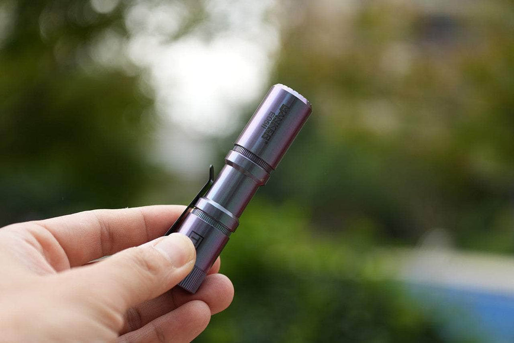 A hand holds the Manker E05 II Ti - Colorful MAO Limited Edition, a compact titanium flashlight with a pocket clip, shining brightly outdoors against lush greenery for perfect everyday carry.