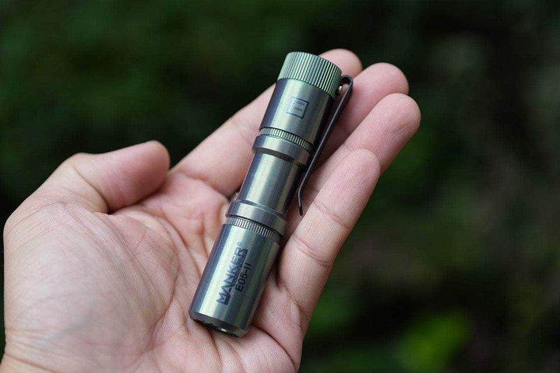 A hand holds the colorful MAO limited edition Manker E05 II Ti flashlight with a clip, shining brightly—ideal for everyday carry—against a blurred outdoor background.
