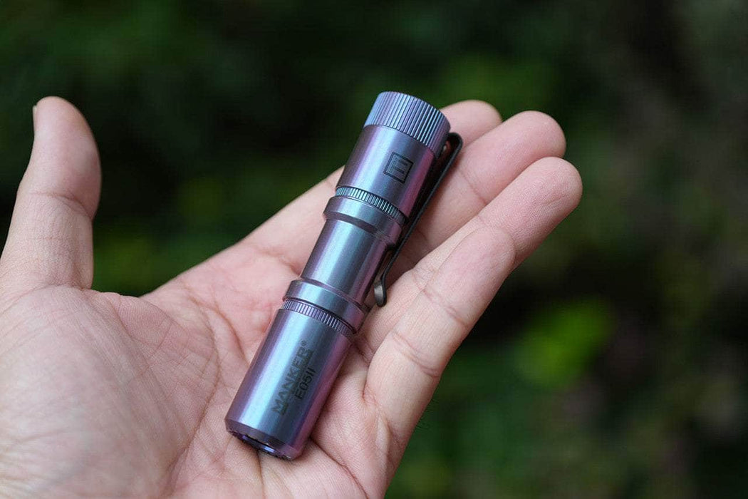 A person holds the Manker E05 II Ti - Colorful MAO Limited Edition, a compact titanium flashlight with 1300 lumens, in their open palm against a blurred outdoor backdrop—an ideal everyday carry accessory.