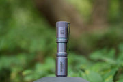 The Manker E05 II Ti - Colorful MAO Limited Edition, a compact titanium flashlight with pocket clip, stands upright on a surface—ideal for everyday carry—set against a blurred green outdoor background.