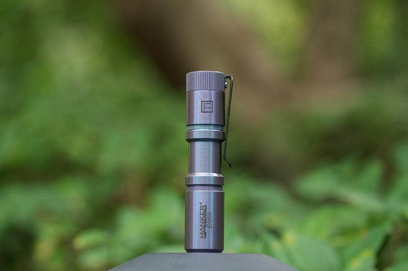 The Manker E05 II Ti - Colorful MAO Limited Edition, a compact titanium flashlight with pocket clip, stands upright on a surface—ideal for everyday carry—set against a blurred green outdoor background.