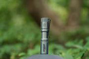 The Manker E05 II Ti - Colorful MAO Limited Edition flashlight stands upright outdoors amid green foliage and a blurred natural background, making it a stylish choice for everyday carry.