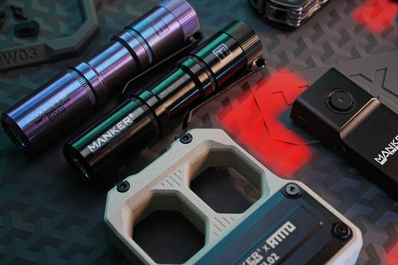 The Manker E05 II Titanium DLC Black flashlight, a compact model with 1300 lumens output, is displayed with another Manker flashlight, tactical gear, and a small black device on a textured black surface highlighted by red light—ideal for everyday carry.