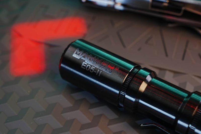 A close-up of a Manker E05 II Titanium DLC Black everyday carry flashlight on a textured surface with red and blue reflections, highlighting its compact design and powerful 1300 lumens output.