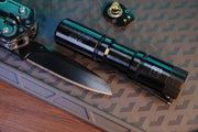 A Manker E05 II Titanium DLC Black flashlight with 1300 lumens sits beside a partially open folding knife on a textured mat, along with a small battery or electronic component.