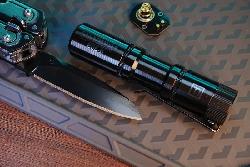 A Manker E05 II Titanium DLC Black flashlight with 1300 lumens sits beside a partially open folding knife on a textured mat, along with a small battery or electronic component.