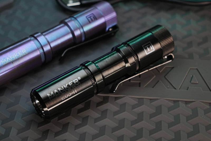 A Manker E05 II Titanium DLC Black everyday carry flashlight rests on a textured surface with a partial "EXA" logo in the background. It delivers up to 1300 lumens for powerful illumination.