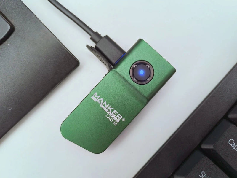 A green Manker LAD Mini Rechargeable Keychain Flashlight with a blue power button is connected to a USB-C charging cable, positioned between a laptop and a keyboard.