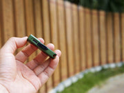 A hand holds the Manker LAD Mini Rechargeable Keychain Flashlight, a compact green EDC light with a visible USB-C port, outdoors near a wooden fence.