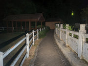 A paved pathway with white fences curves past a gazebo and shrubs at night, illuminated by the beam of a Manker LAD Mini Rechargeable Keychain Flashlight.