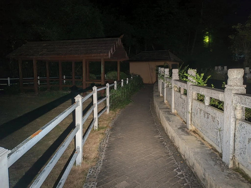 A paved pathway with white fences curves past a gazebo and shrubs at night, illuminated by the beam of a Manker LAD Mini Rechargeable Keychain Flashlight.