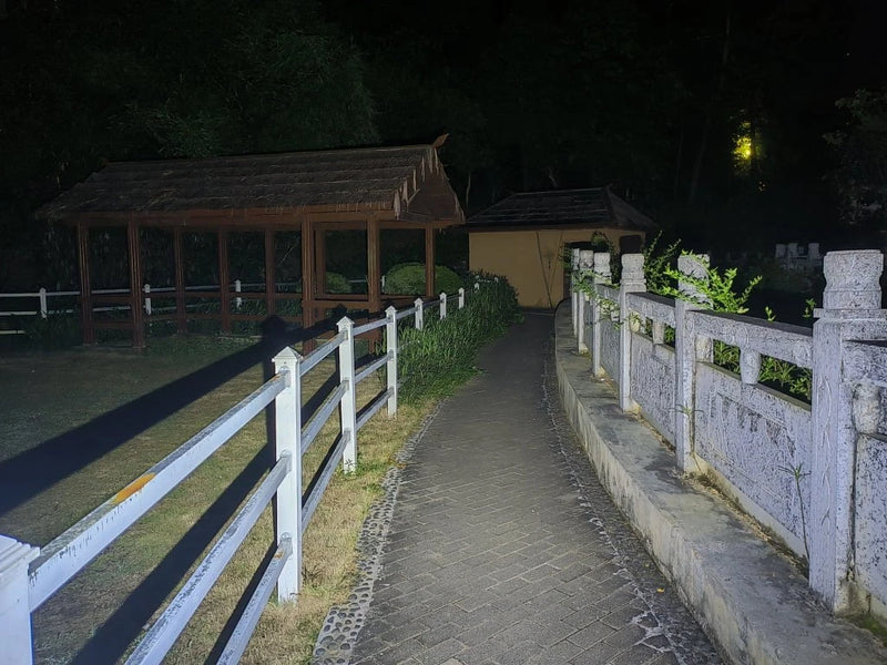 A paved pathway between a white fence and stone railing leads to illuminated wooden and tan structures at night—ideal for navigation using the Manker LAD Mini Rechargeable Keychain Flashlight with USB-C charging.