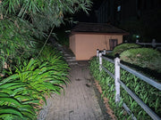A paved path at night is lit by the Manker LAD Mini Rechargeable Keychain Flashlight, bordered by green plants and a white fence, leading to a small shed with a dark roof and shuttered window.