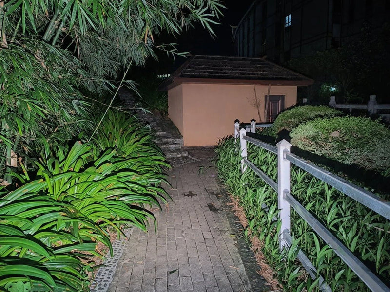 A paved path at night is lit by the Manker LAD Mini Rechargeable Keychain Flashlight, bordered by green plants and a white fence, leading to a small shed with a dark roof and shuttered window.