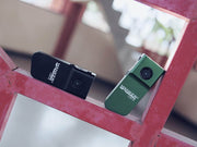 Two Manker LAD Mini Rechargeable Keychain Flashlights, one black and one green, are placed on an indoor red geometric metal structure.