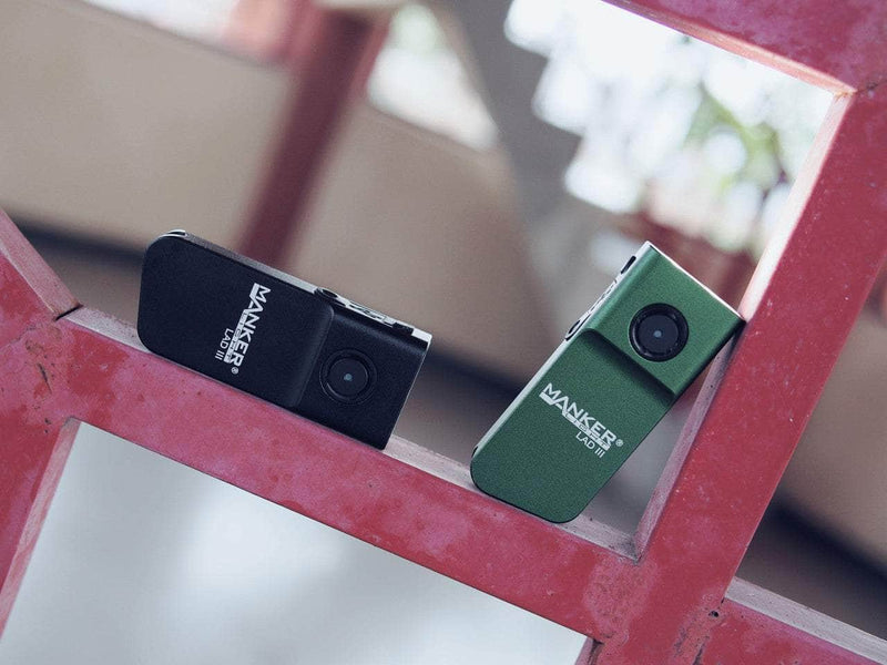 Two Manker LAD Mini Rechargeable Keychain Flashlights, one black and one green, are placed on an indoor red geometric metal structure.