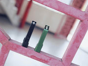 Two rectangular Manker LAD Mini Rechargeable Keychain Flashlights, one black and one green, stand upright on a pink metal grid with a blurred indoor background, showcasing their sleek design and convenient USB-C charging.