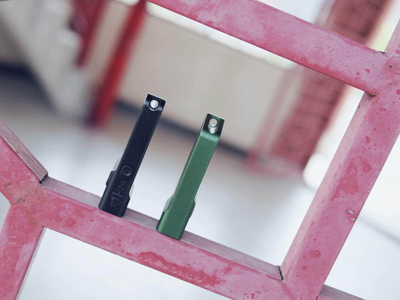 Two rectangular Manker LAD Mini Rechargeable Keychain Flashlights, one black and one green, stand upright on a pink metal grid with a blurred indoor background, showcasing their sleek design and convenient USB-C charging.