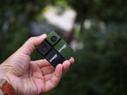 A hand holds two compact LAVAKERS cameras—one black and one green—alongside a Manker LAD Mini Rechargeable Keychain Flashlight, set against a blurred outdoor background.