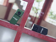 Two Manker LAD Mini Rechargeable Keychain Flashlights, one green and one black, sit on a red metal grid with a blurred outdoor background. These compact keychain lights feature USB-C charging for convenient everyday carry.