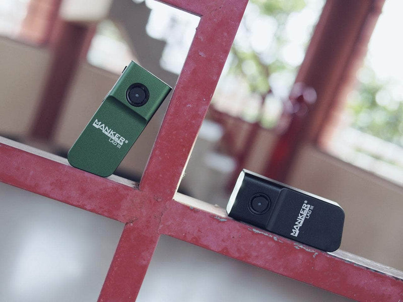 Two Manker LAD Mini Rechargeable Keychain Flashlights, one green and one black, sit on a red metal grid with a blurred outdoor background. These compact keychain lights feature USB-C charging for convenient everyday carry.