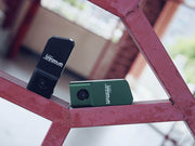 Two Manker LAD Mini Rechargeable Keychain Flashlights, one black and one green, are displayed on a red metal structure outdoors.