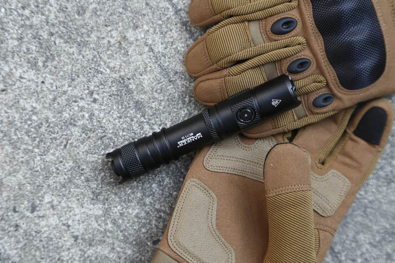 A Manker MC11 III USB-C rechargeable tactical flashlight rests on a pair of brown and tan gloves atop a rough concrete surface.