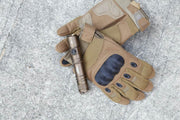 A pair of brown tactical gloves with black knuckle guards lies on concrete, topped by a bronze Manker MC11 III USB-C Rechargeable Tactical Flashlight.